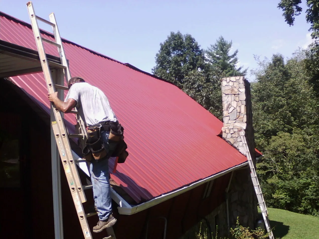Roofer on a ladder beside a red metal roof and stone chimney — Soffit & Fascia Repair by Roofing Brothers in Fairview Heights